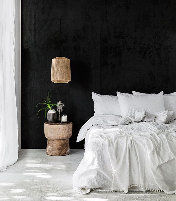 a chic black and white bedroom with black walls, a white floor, white textiles and a wooden lamp and nightstand