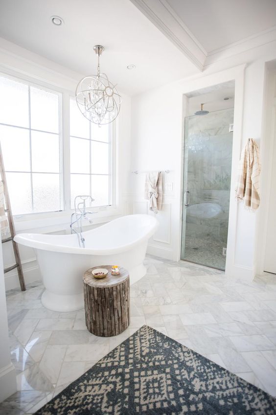 A gorgeous transitional space with marble tiles, a wooden stool, a printed rug, a catchy chandelier and a vintage inspired tub