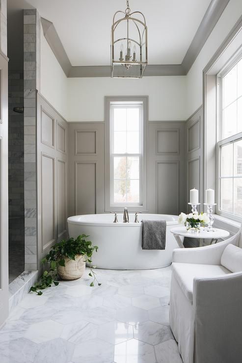 a gorgeously elegant bathroom done with light grey paneling, marble hex tiles on the floor, white furniture and a contemporary oval tub
