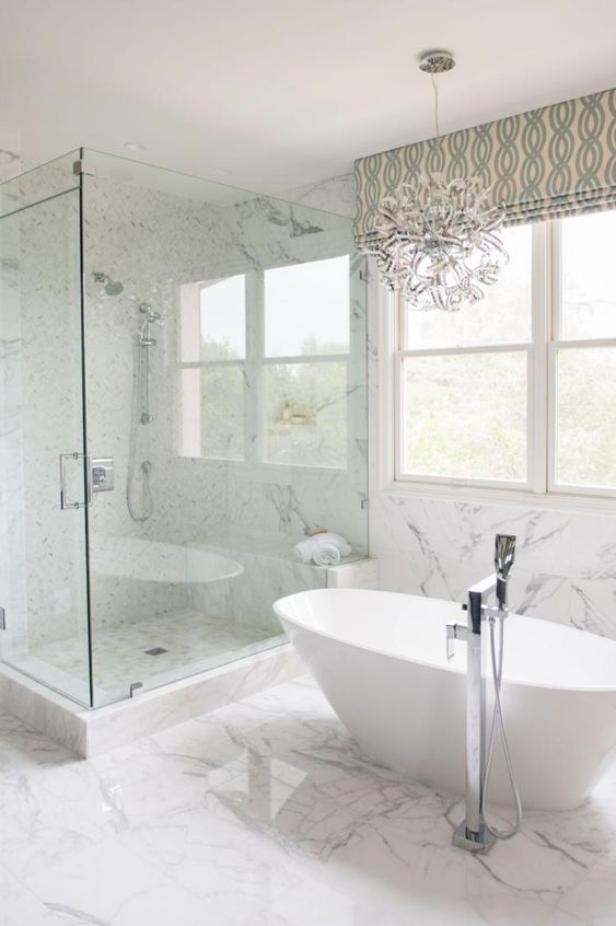 a luxurious transitional space with a fantastic chandelier, an oval tub, marble tiles and printed shades