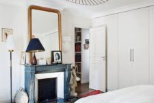 a neutral Parisian bedroom with a blue non-workign fireplace, a statement lamp, a chic mirror and an antique wooden chair