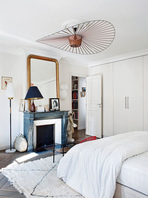 A neutral Parisian bedroom with a blue non workign fireplace, a statement lamp, a chic mirror and an antique wooden chair