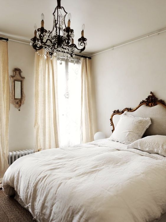 a neutral Parisian bedroom with a refined bed, a crystal chandelier, a mirror and flowy draperies