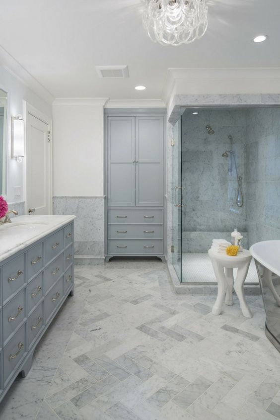 a relaxing bathroom done in greys and neutrals, with a chic chandelier, stylish vintage storage units and marble tiles