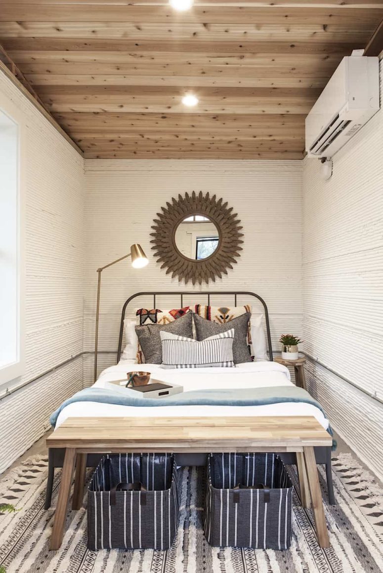 The bedroom is small yet cozy, with a metal bed, a wooden bench and a statement mirror plus a floor lamp