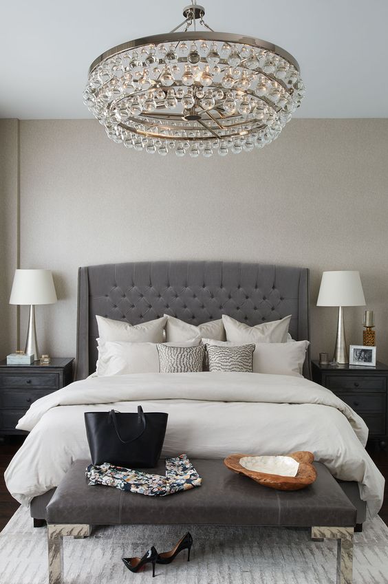 an elegant charcoal grey tufted wingback headboard, a crystal chandelier and a leather bench make up a glam modern space