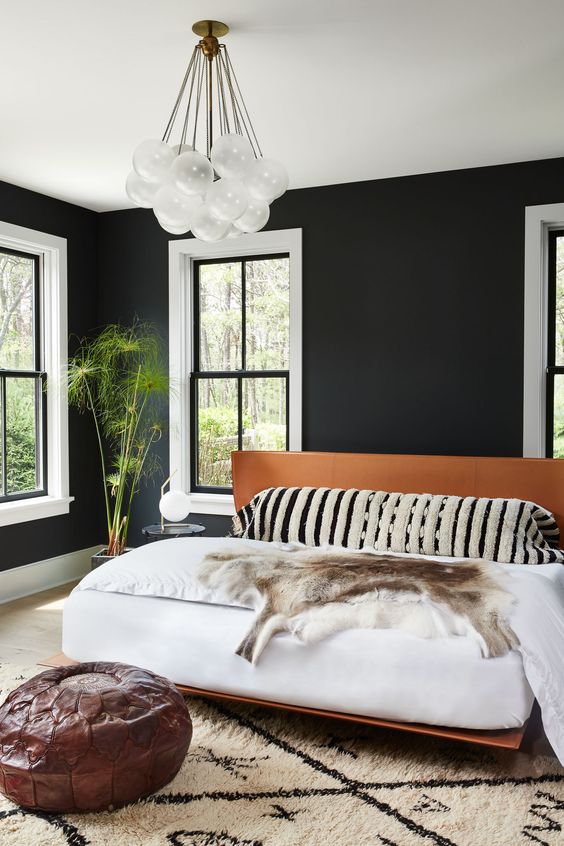 A stylish mid century modern bedroom with black walls, a creative chandelier, a leather bed and white windows