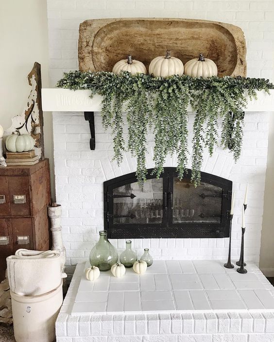 a mantel decorated with cascading greenery, three white pumpkins and a dough bowl