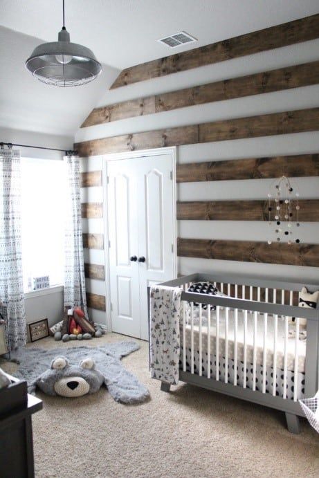 a chic woodland nursery with a striped stained wood wall, layered rugs, a grey vintage crib, a pendant lamp and lace curtains