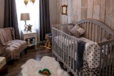 a cool rustic nursery with a reclaimed wood wall, a large grey crib, a fluffy rug, dark curtains and a vintage chandelier