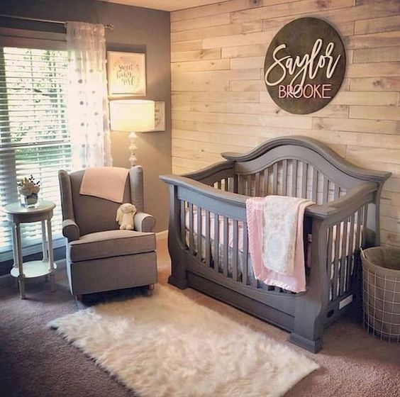 a cozy small farmhouse space with a reclaimed wooden wall, a grey crib and chair, a fluffy rug and shades on the window