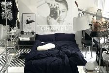 a fashion-inspired bedroom with double-height ceiling, a bed with black bedding, glass shelving units, a vanity and some art