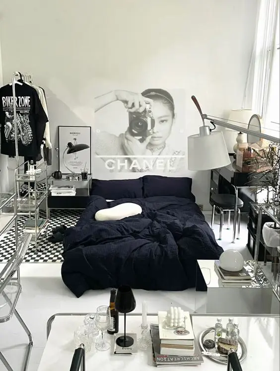 A fashion inspired bedroom with double height ceiling, a bed with black bedding, glass shelving units, a vanity and some art