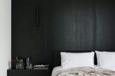 a minimal monochromatic bedroom with a black accent wall, a black bed and neutral bedding, a floating nightstand and a lamp