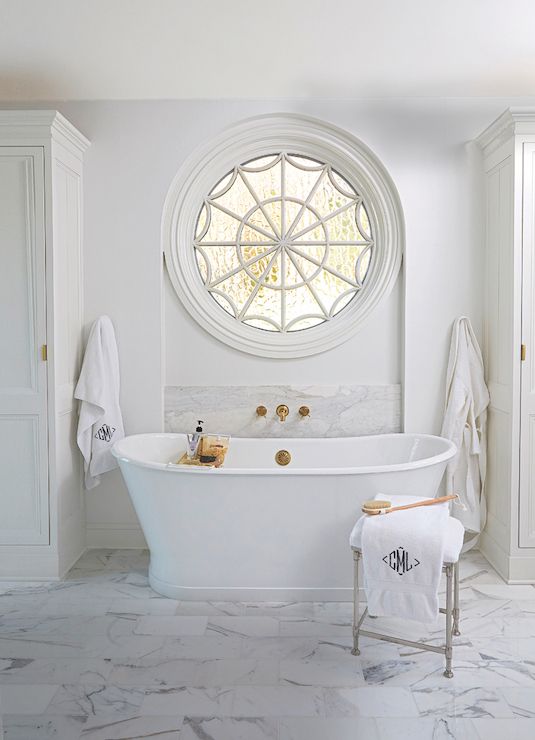 a neutral farmhouse bathroom done with marble tiles, a chic tub and a porthole window next to the tub