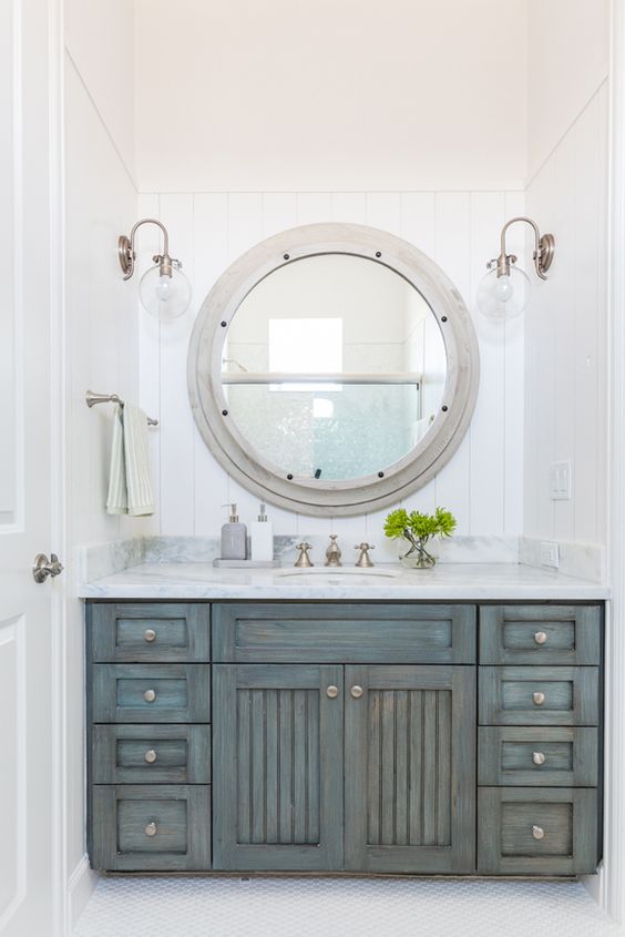 a shabby chic coastal bathroom with an ged wood vanity and a large porthole window to the shower