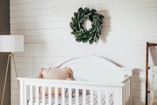 a simple nursery done with a white shiplap wall, a white vintage crib, a sphere chandelier and a faux greenery wreath