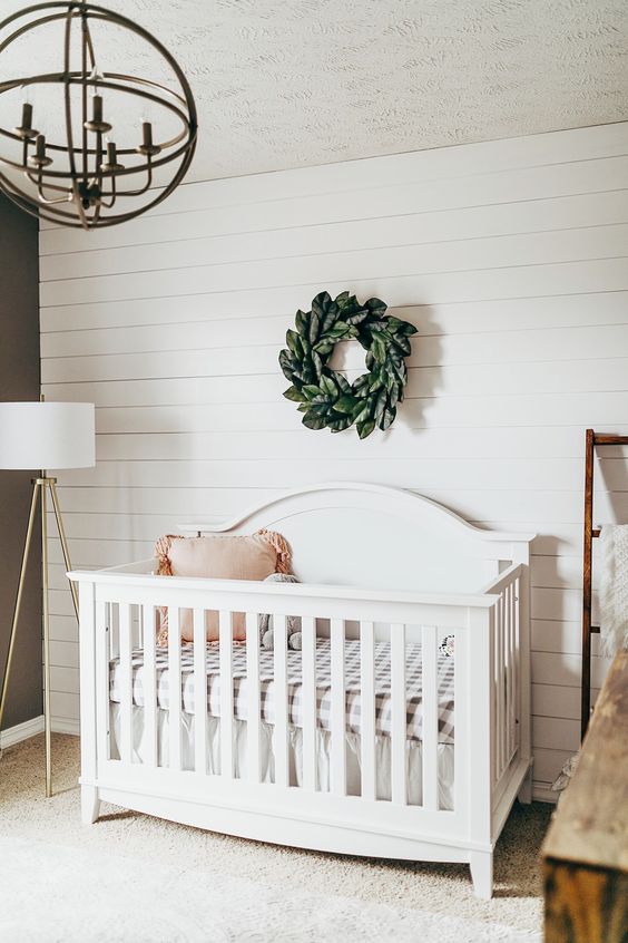 a simple nursery done with a white shiplap wall, a white vintage crib, a sphere chandelier and a faux greenery wreath