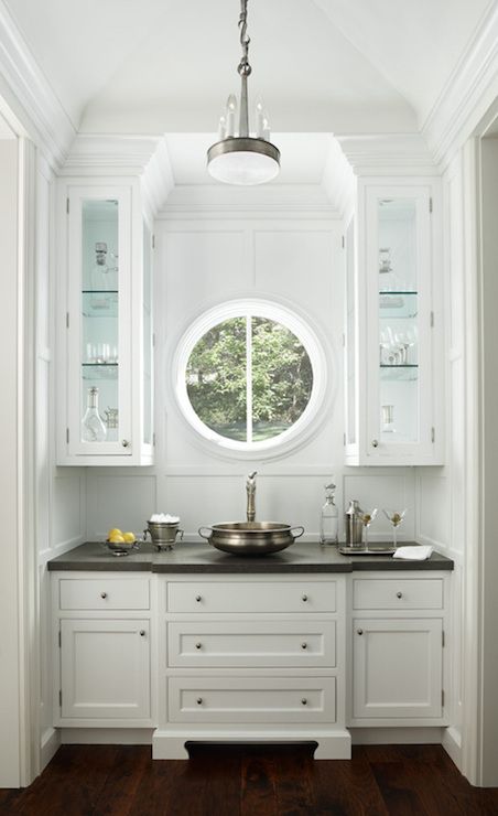 a small home bar with traditional white cabinets and a small porthole window to make the space unique