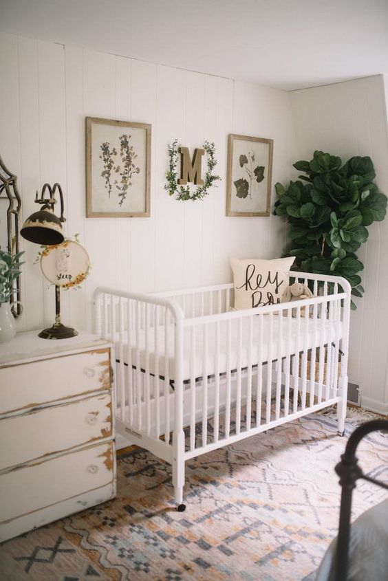 a vintage farmhouse nursery with a white crib, a shabby chic dresser, a printed rug and greenery