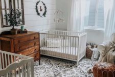 a welcoming shared farmhouse nursery with a printed rug, a leather ottoman, a stained dresser, a bead chandelier and some greenery
