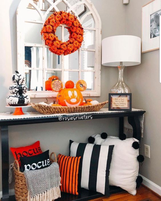 a bright Halloween entryway console with black, white and orange pillows, a Jack-o-lantern, a pumpkin wreath and a stack of white pumpkins
