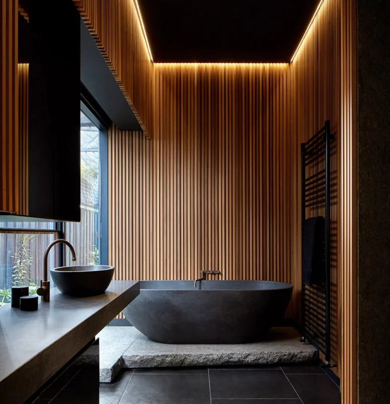 The wonderful moody bathroom is done with a stone tub on a platform, built-in lights over it, a stone vanity and some minimalist textures