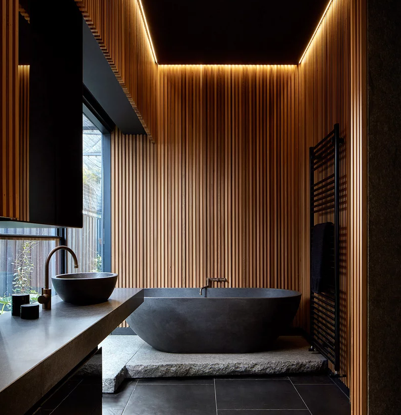 The wonderful moody bathroom is done with a stone tub on a platform, built in lights over it, a stone vanity and some minimalist textures
