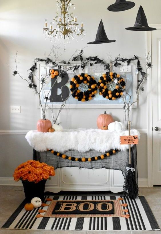 A Halloween entryway console with a printed rug, a bold floral arrangement, pompom wreaths, branches with spiders and witches' hats