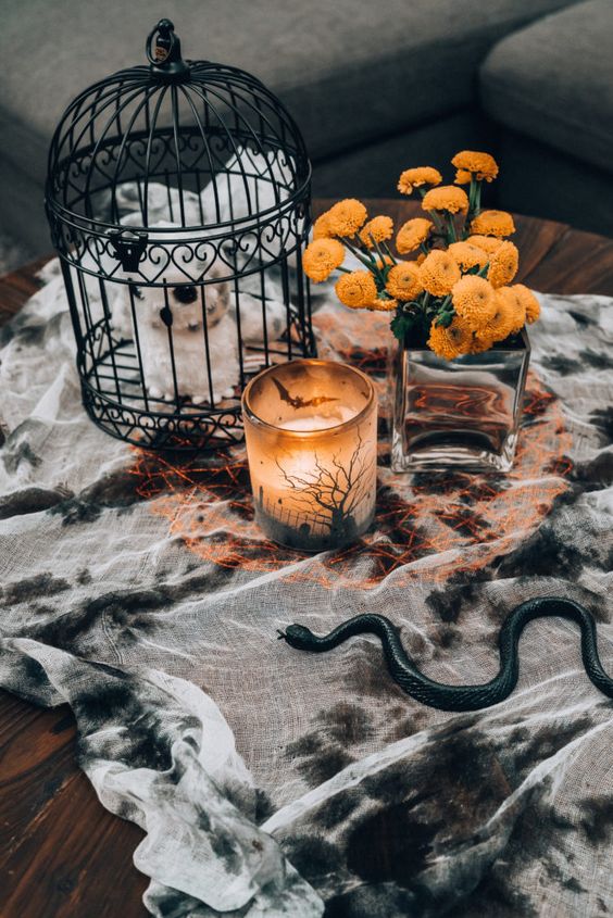 stylish Halloween coffee table decor with a splatted tablecloth, a cage with a plush, a candleholder and bold blooms in a vase