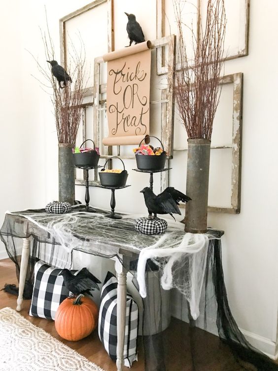 A Halloween entryway with spiderweb, checked pillows, a pumpkin, blackbirds, branches in buckets and empty frames