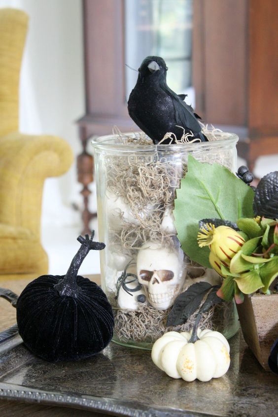 a glass jar with hay and skulls, some faux pumpkins, greenery and a blackbird on top for Halloween