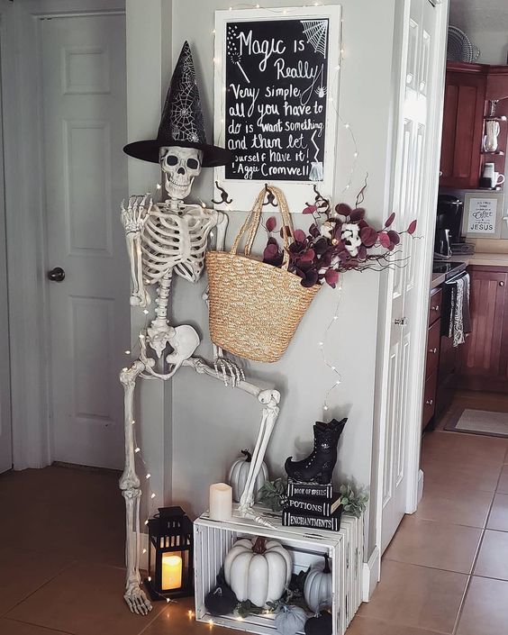Halloween entryway decor with a skeleton, lights, a candle lantern, a create with pumpkins and a straw bag with dark blooms