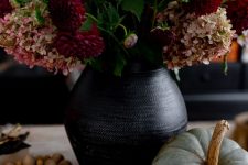 a Halloween centerpiece of a black vase, dark blooms, some pumpkins including painted ones and wooden beads