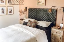 a cozy boho bedroom with a dusty pink wall, a bed with a black woven leather headboard, monochromatic bedding, brass lamps and some artwork