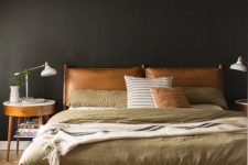 a moody mid-century modern bedroom with black walls, a bed with an amber leather headboard and muted color bedding and a nightstand