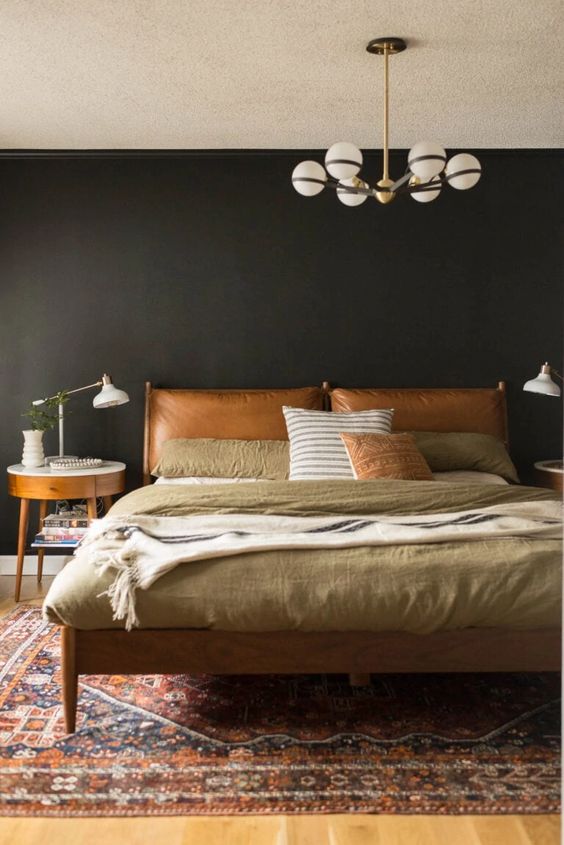 A moody mid century modern bedroom with black walls, a bed with an amber leather headboard and muted color bedding and a nightstand