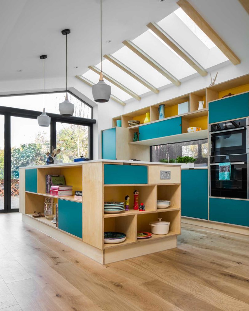 The kitchen is done in light-colored plywood and with teal panels, concrete pendant lamps and a window backsplash