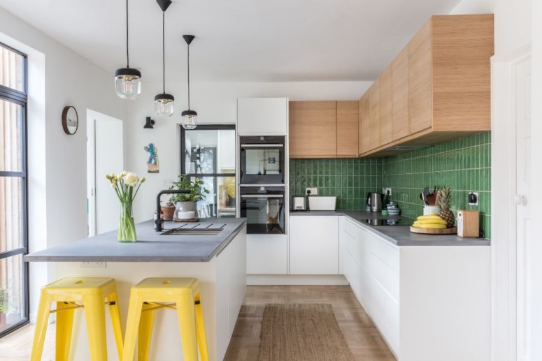 The kitchen is done with sleek cabinets and a green tile backsplash, a kitchen island with a concrete countertop and bright yellow stools