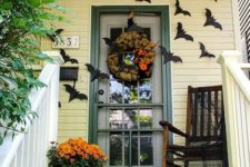 a Halloween porch with black bats on the walls, a mesh wreath, orange blooms, a carved pumpkin lantern