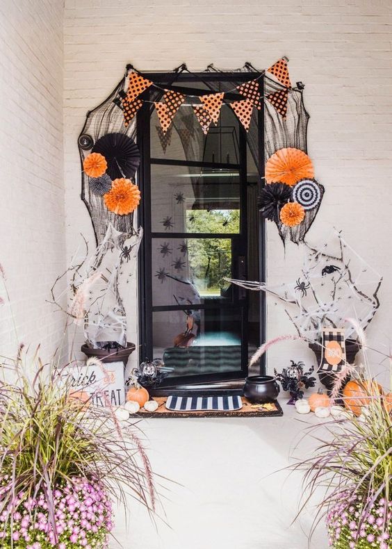 a bold and whimsical Halloween porch with paper fans, buntings, orange and white pumpkins, spiders and branches