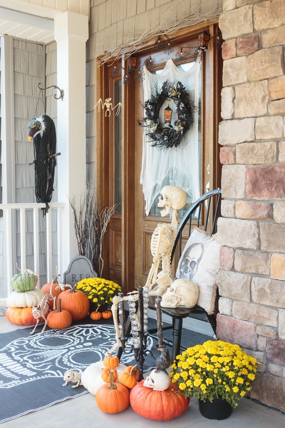 a bright Halloween porch with a black feather wreath, a skeleton and skulls, bright fall blooms and natural pumpkins
