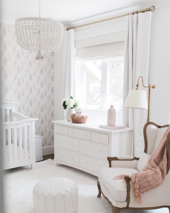 a chic neutral nursery with a bead chandelier, a vintage chair and crib, a dresser, white rugs and a Moroccan pouf