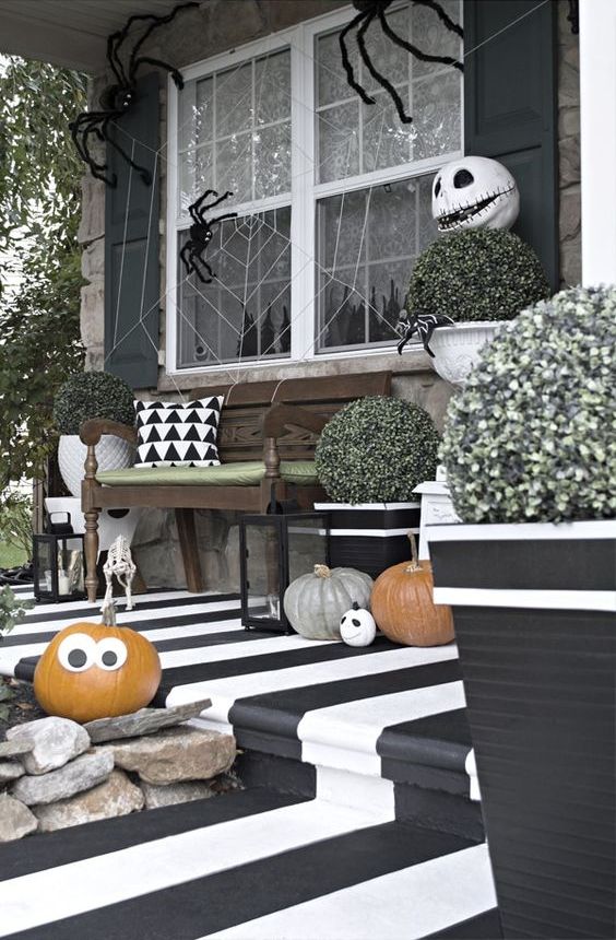 a fun black and white Halloween porch with a striped floor, natural pumpkins with faces, greenery, a spiderweb with spiders