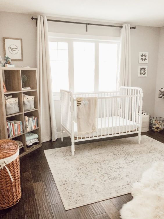A light filled neutral nursery with a crib, a wooden shelving unit, a printed rug, baskets and neutral curtains