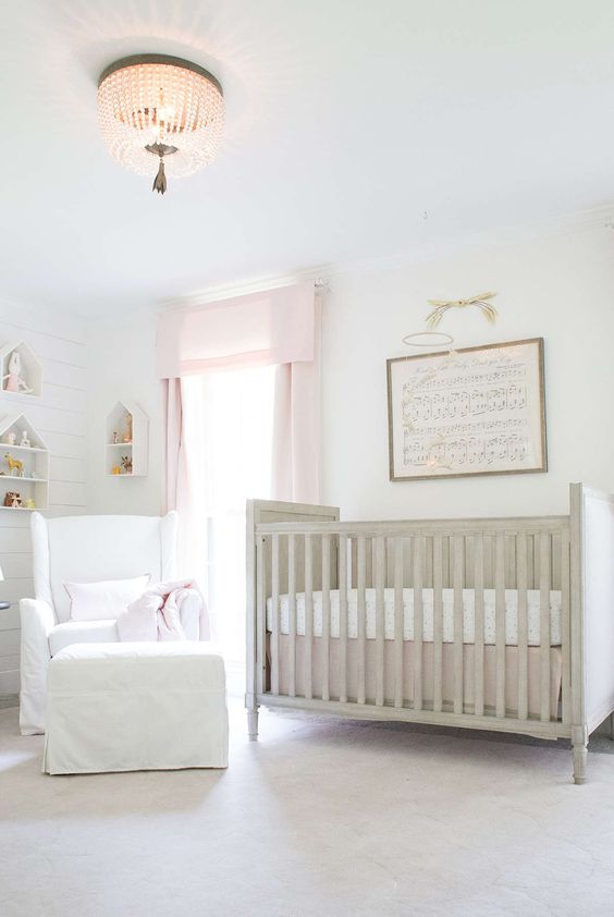 A neutral nursery softened with pinnk curtains, a vintge crib, a white chair with a footrest and house shaped shelves