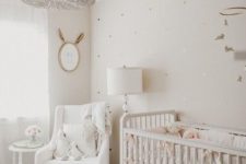 a neutral nursery with a beaded chandelier, a vintage crib, a white chair with a footrest, a mirror and a mobile