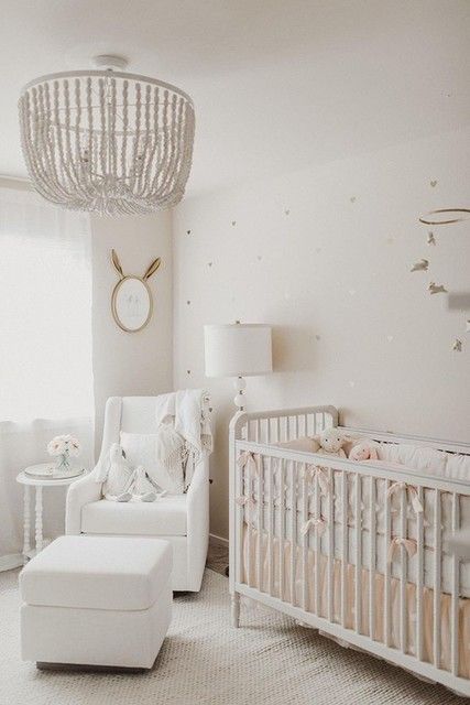 a neutral nursery with a beaded chandelier, a vintage crib, a white chair with a footrest, a mirror and a mobile