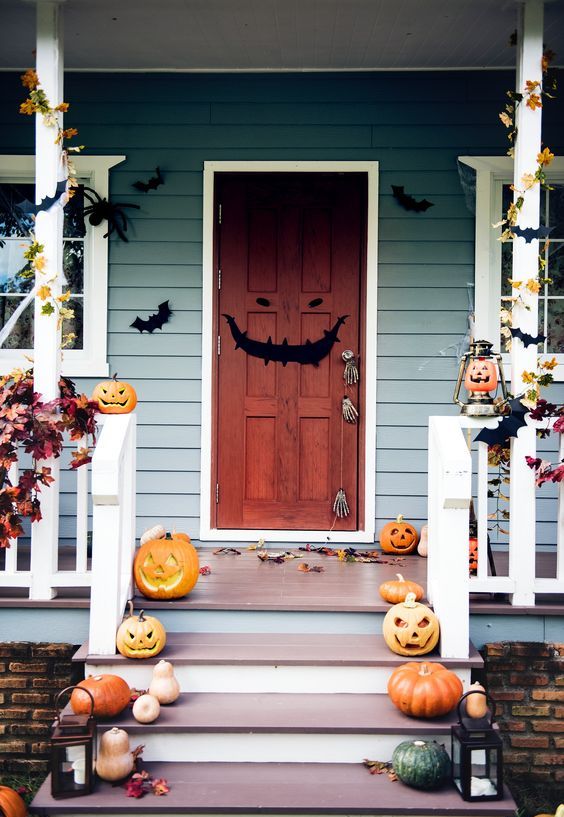 a whimsical Halloween porch with carved pumpkins on steps, candle lanterns, paper bats, fall leaves and a grin door