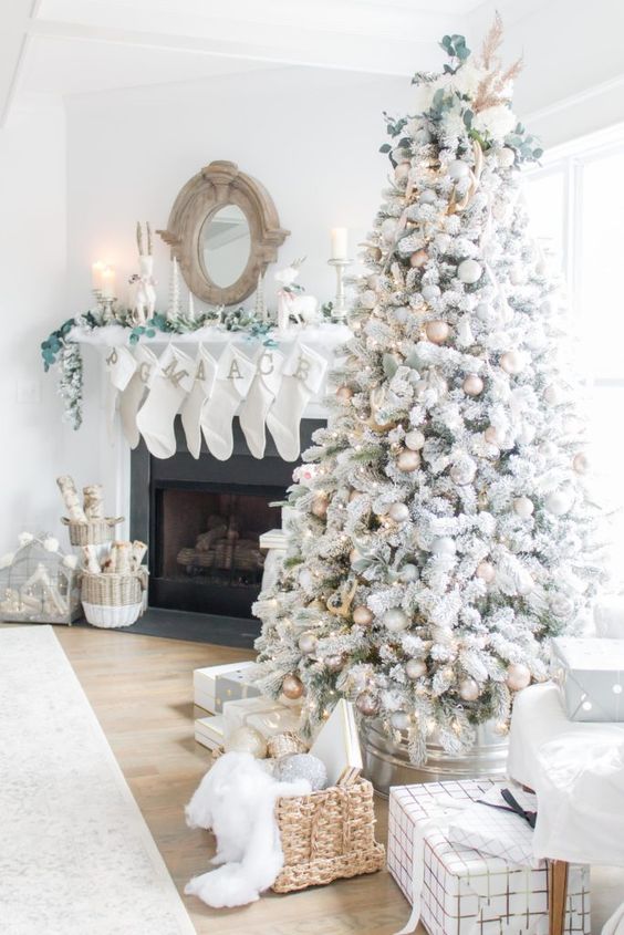 a neutral living room with a flocked Christmas tree with metallic ornaments,  a basket with gifts, white stockings with monogram, greenery and baskets with firewood